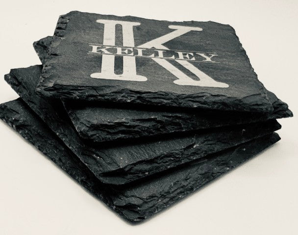 A side view four stacked slate black square coasters with an engraved design. The top coaster features a large, bold letter monogram and the last name horizontally across the middle of the monogram. The engraving is a lighter shade, creating contrast against the dark background of the coaster.
