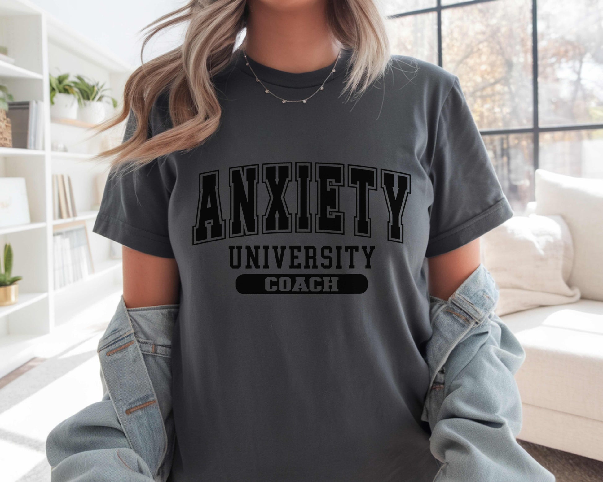 Person wearing a gray t-shirt with 'ANXIETY UNIVERSITY COACH' text in a living room setting.