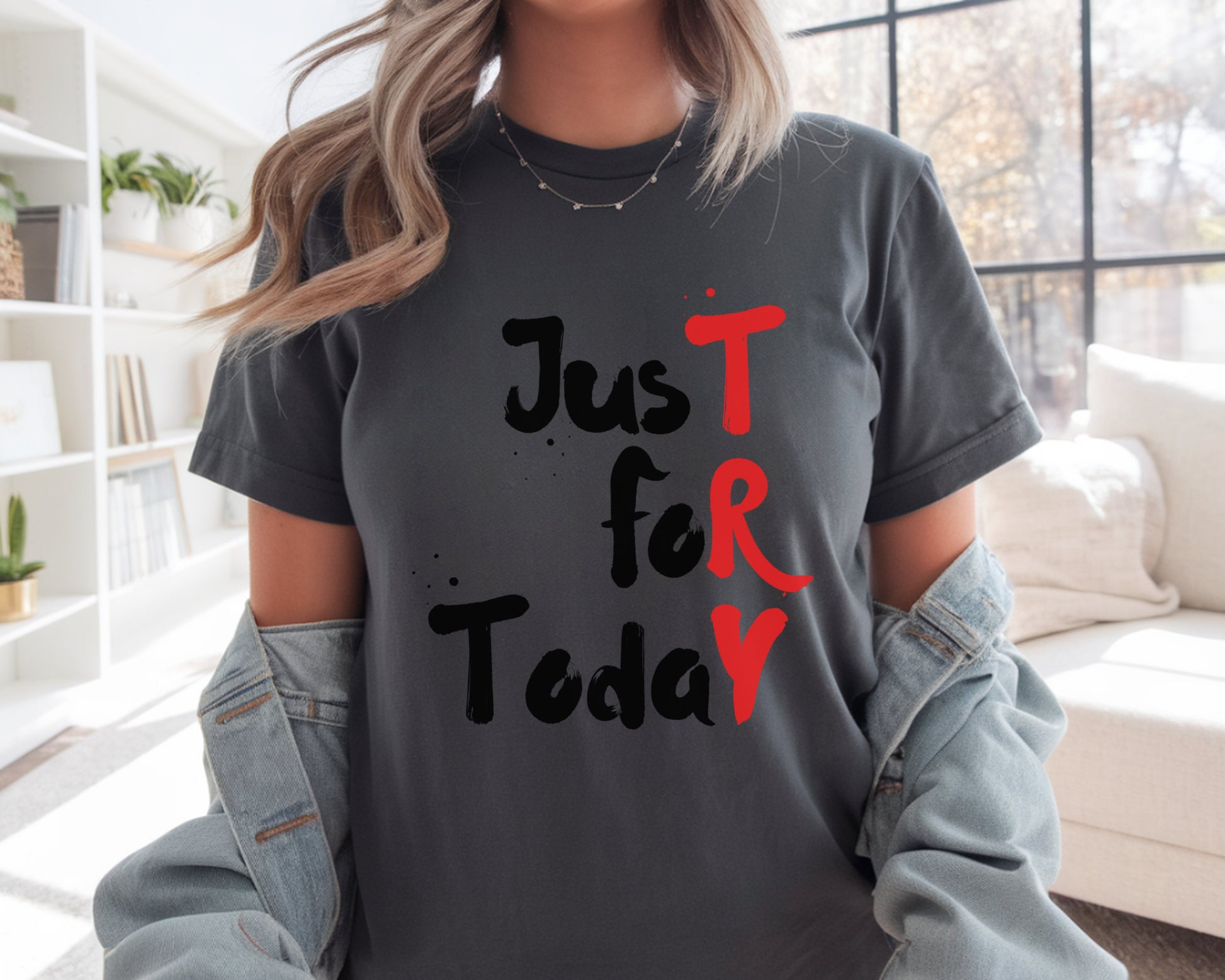 Person wearing a gray t-shirt with 'Just For Today' text in black and red, sitting in a room with a bookshelf and window.