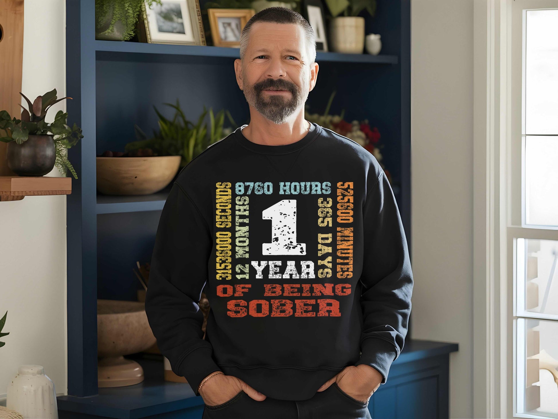 Man wearing a black sweatshirt with '1 year sober', standing in a room with a blue bookshelf and plants.