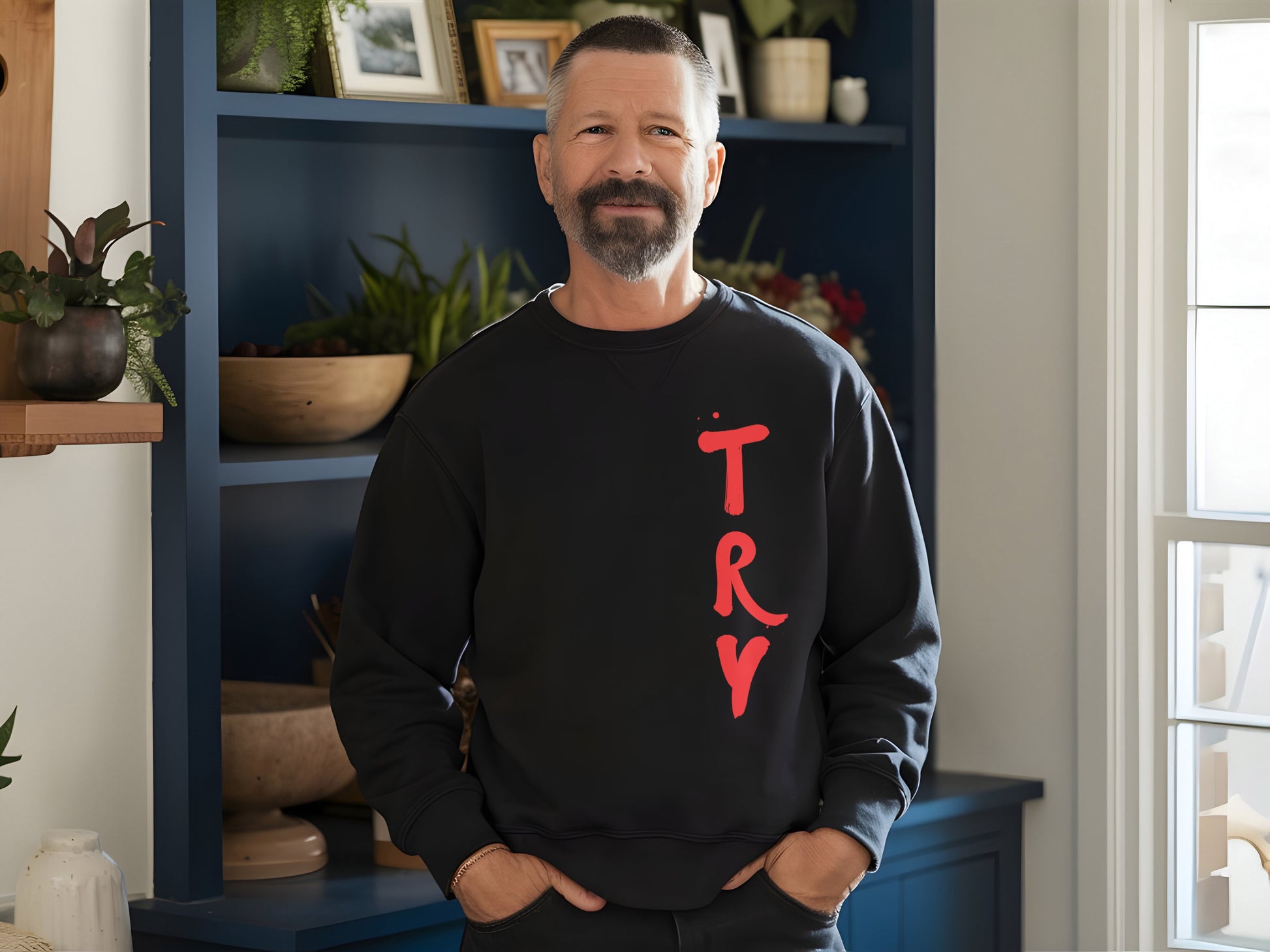 Man wearing a black sweatshirt with 'TRY' in red letters, standing in a room with plants and shelves.