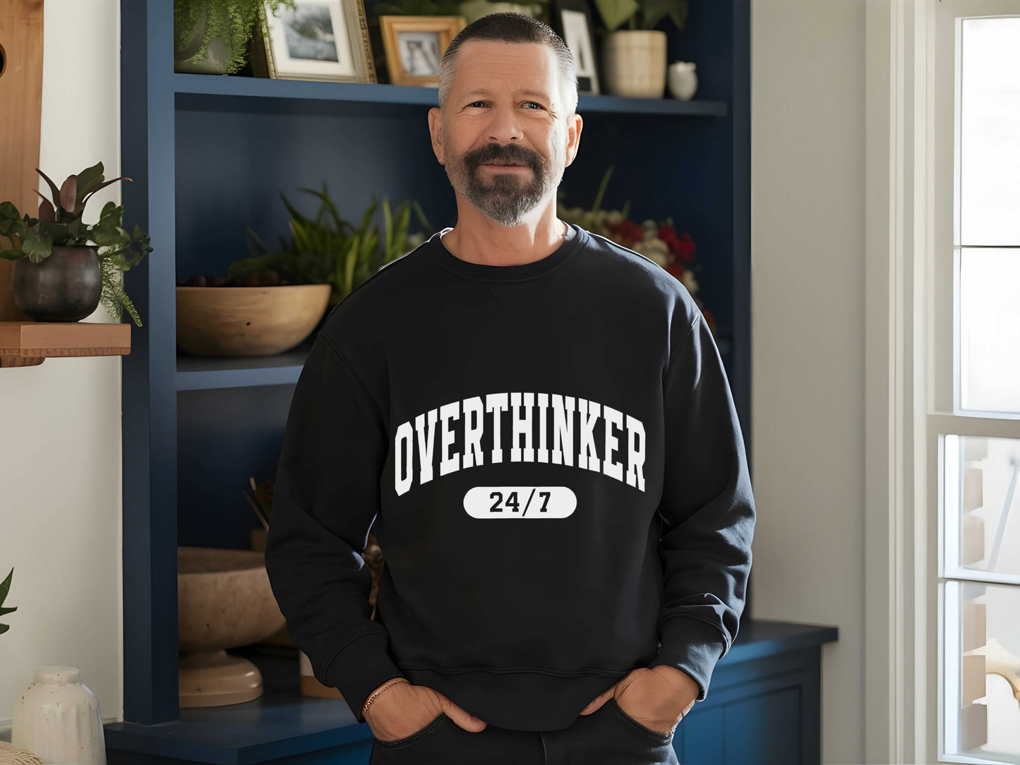 Man wearing a black sweatshirt with 'OVER THINKER 24/7' text in a room with plants and shelves.