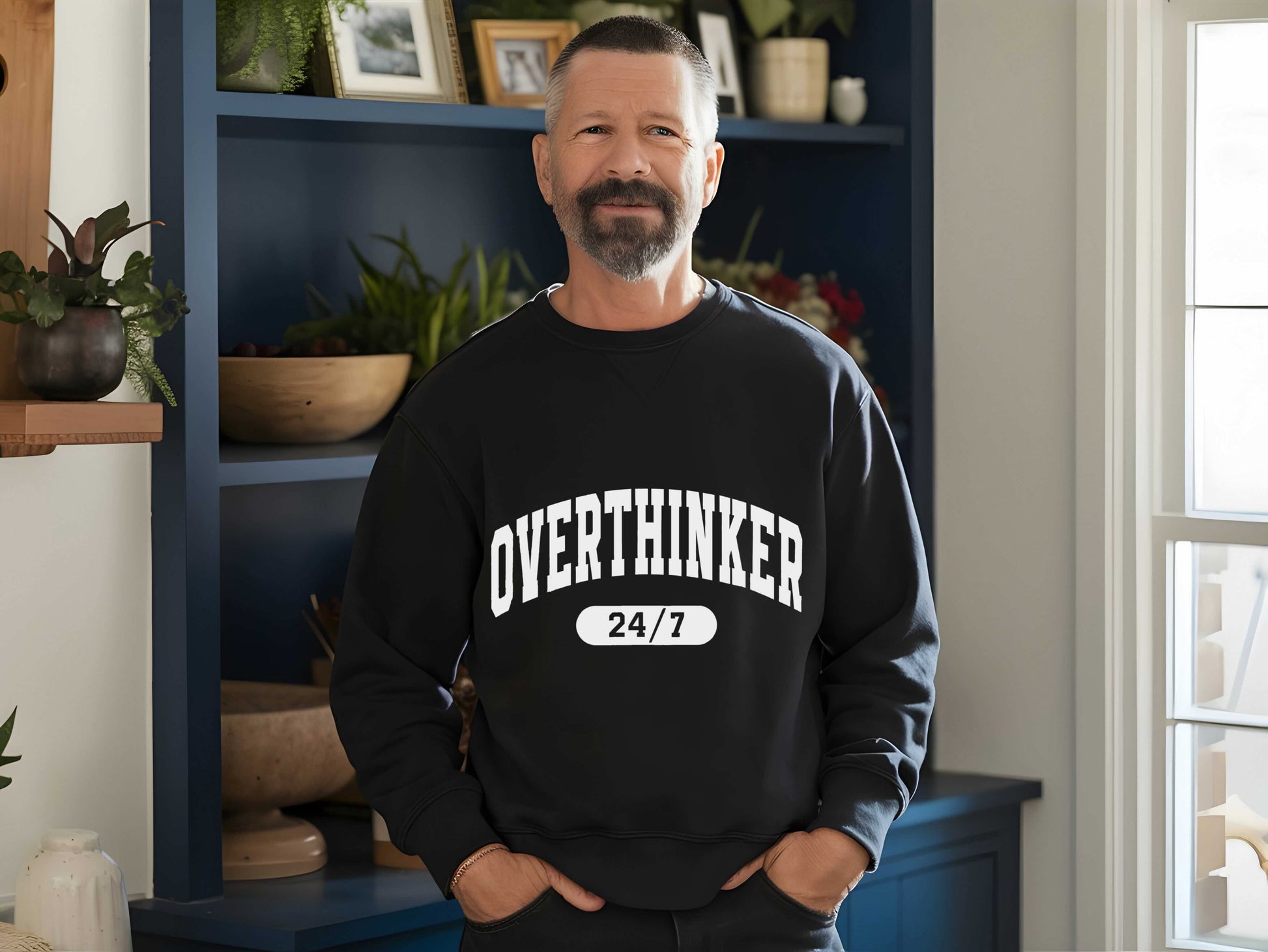 Man wearing a black sweatshirt with 'OVER THINKER 24/7' text in a room with plants and shelves.