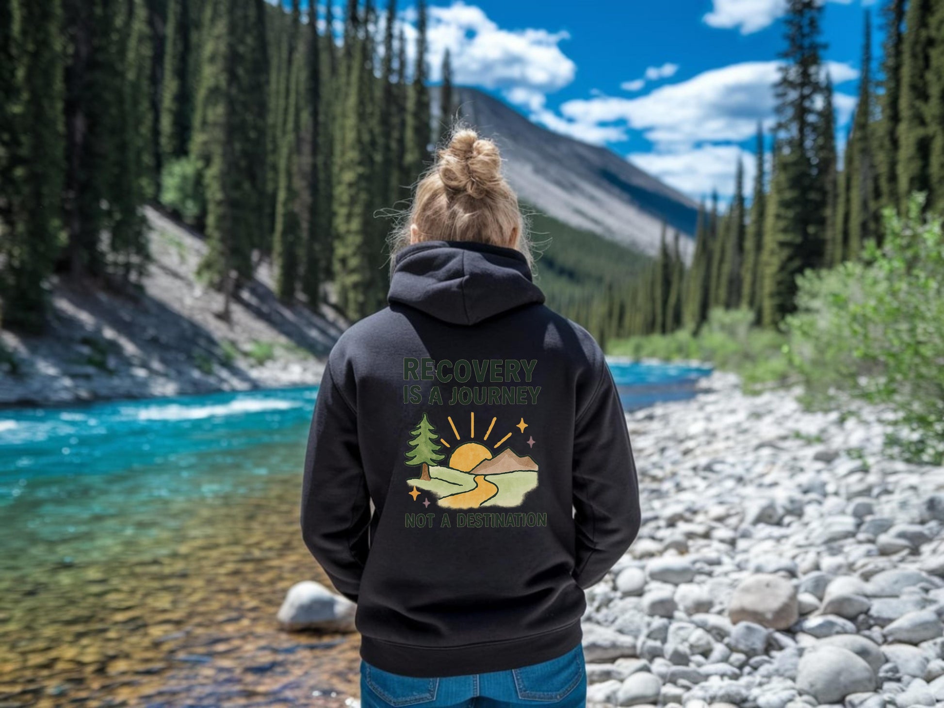 Person wearing a black hoodie with 'Recovery is a Journey, Not a Destination' and a nature-themed design by a river and forest.