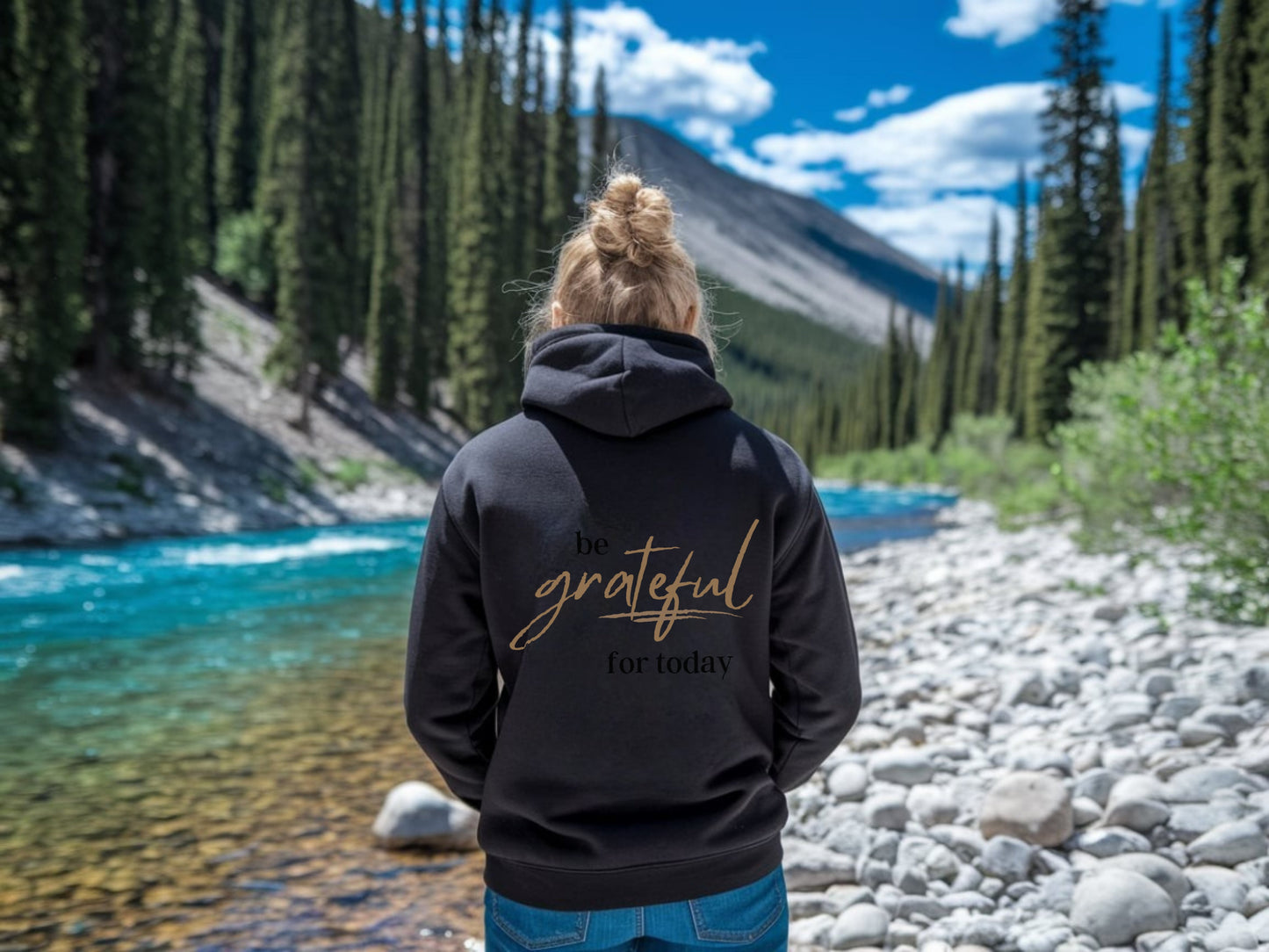 Person wearing a black hoodie with 'Be grateful for today' text in a natural setting with a river and trees.