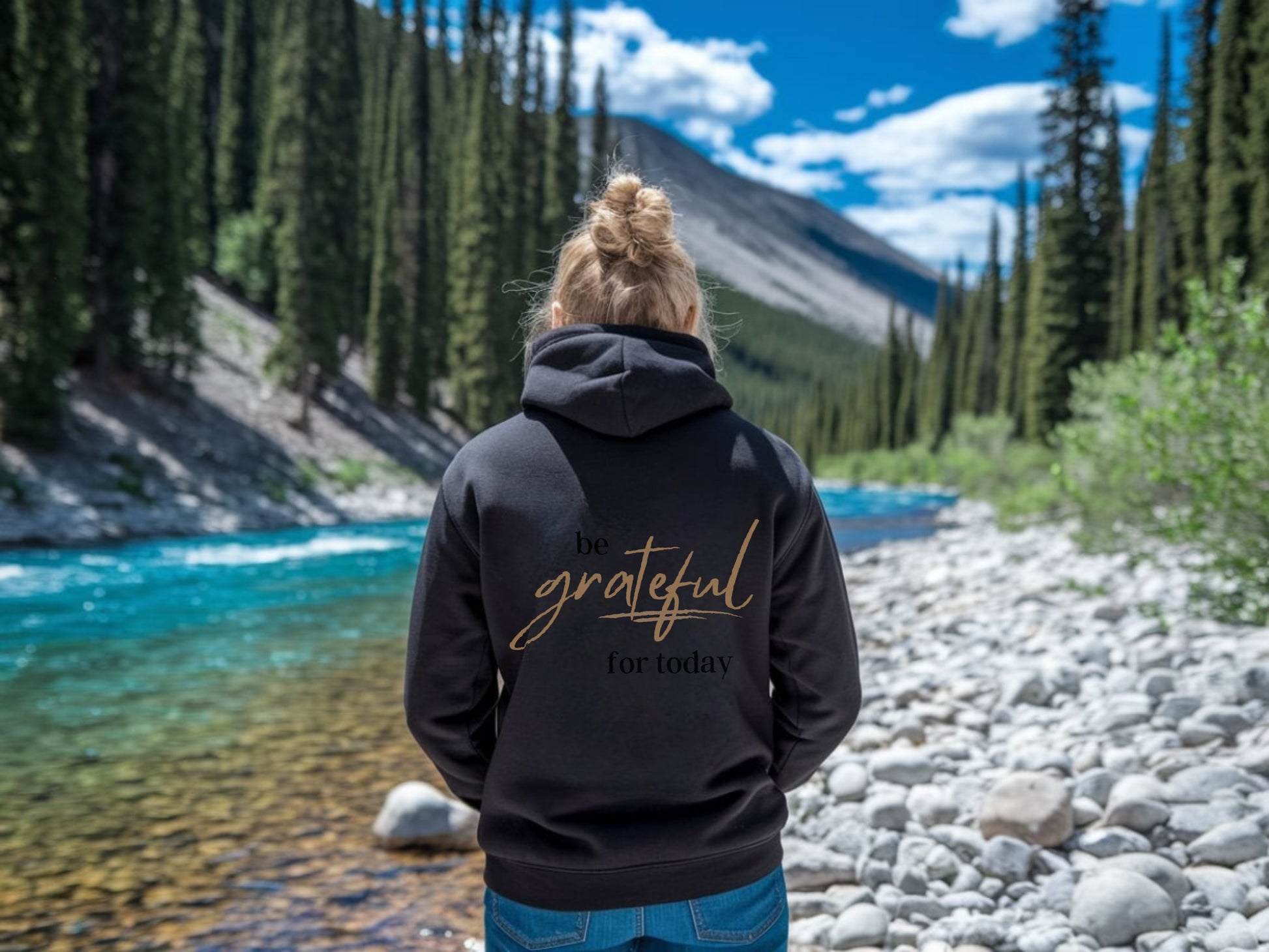 Person wearing a black hoodie with 'Be grateful for today' text in a natural setting with a river and trees.