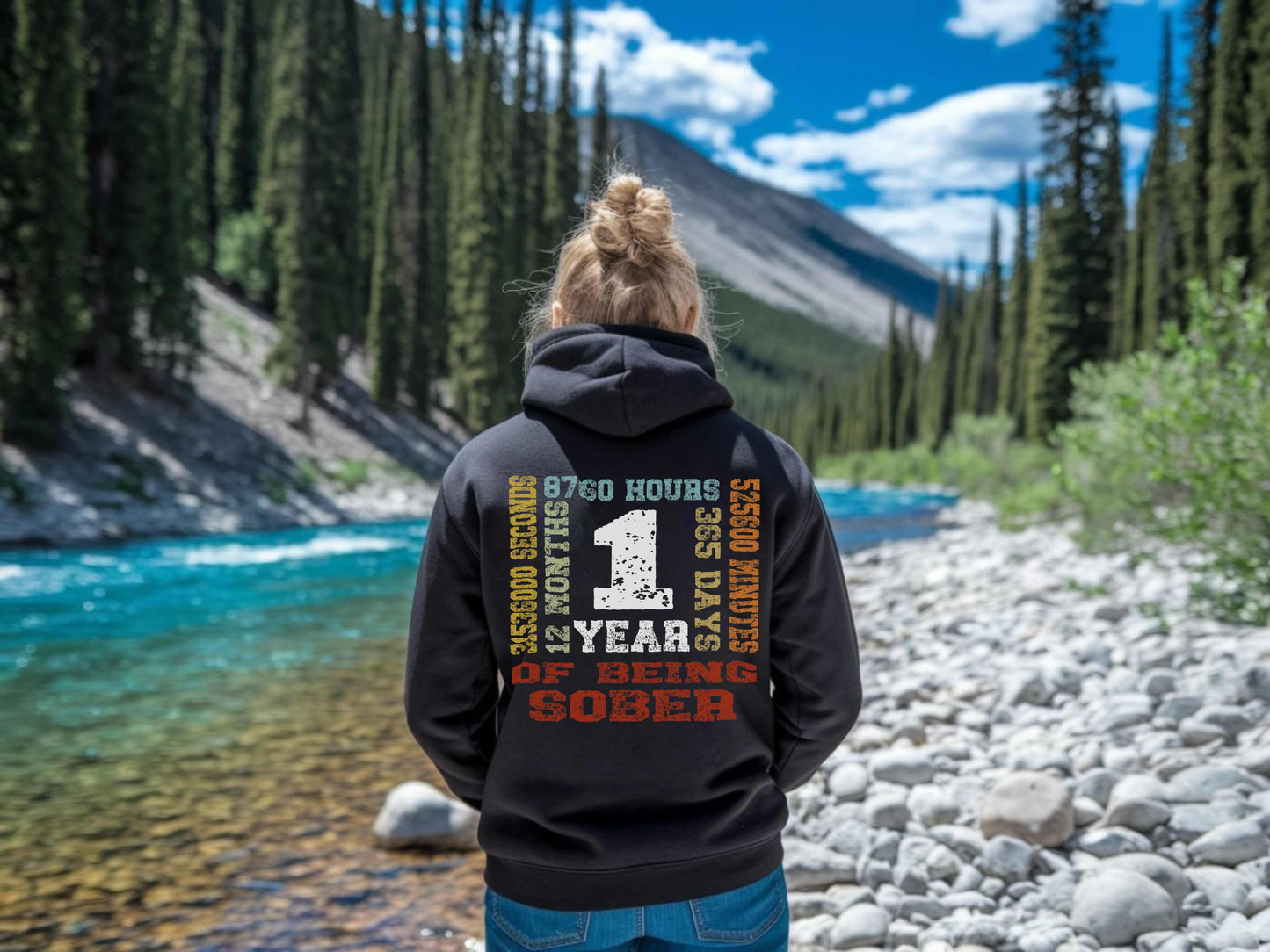 Person wearing a hoodie with '1 year sober' on the back, standing by a river with trees in the background
