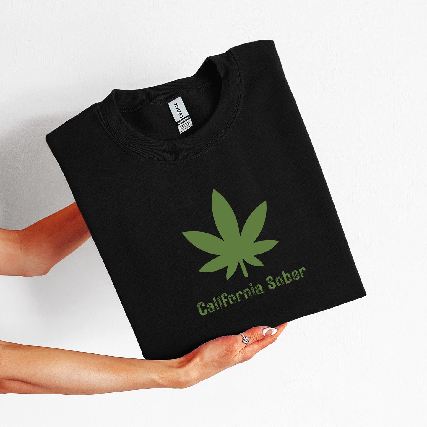Black t-shirt with green cannabis leaf and 'California Sobriety' text held by a person on a white background