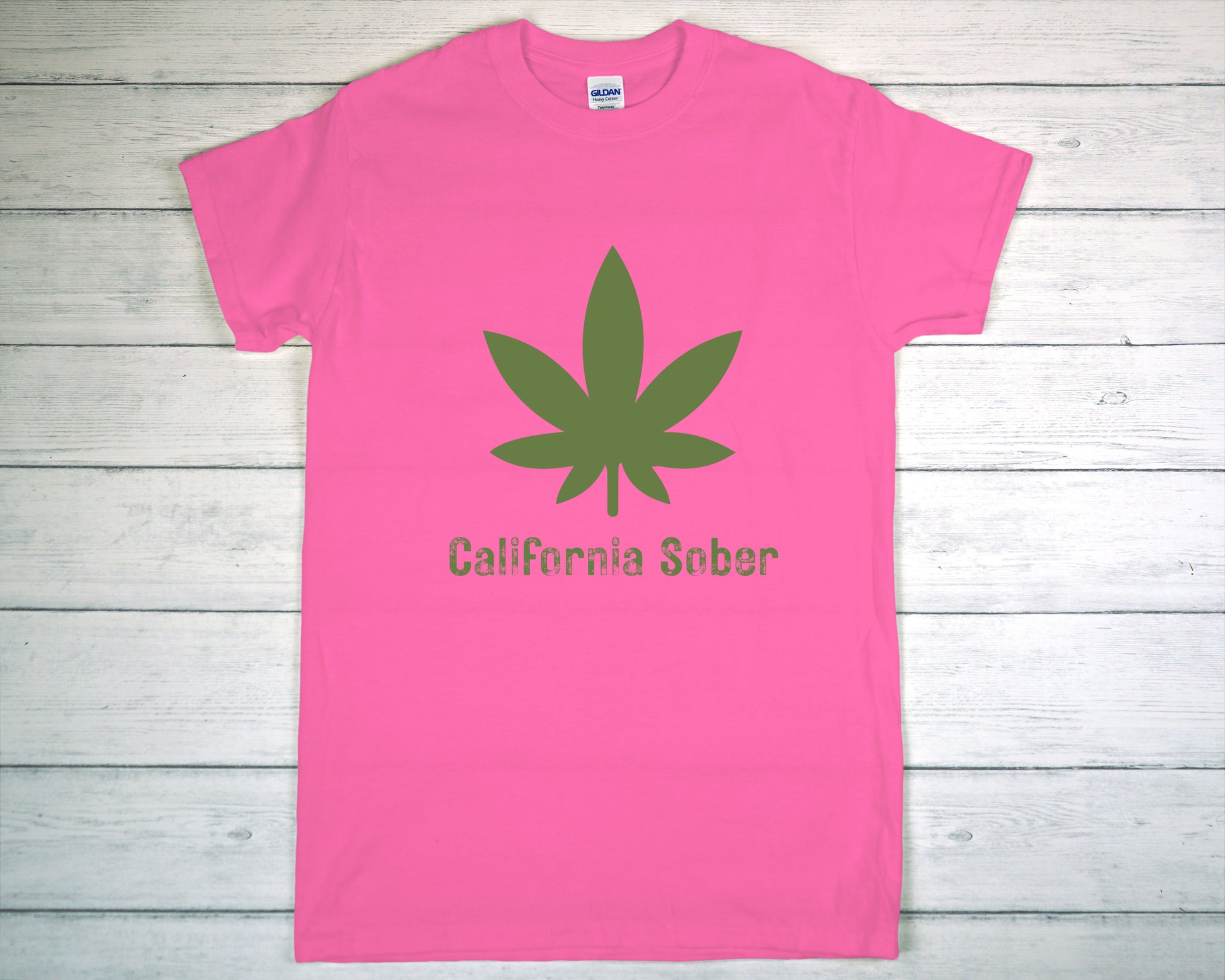 Pink t-shirt with green cannabis leaf and 'California Sober' text on a wooden surface