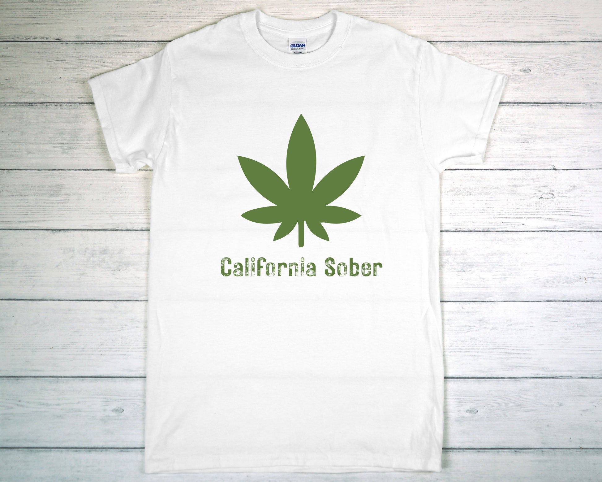 White t-shirt with green cannabis leaf and 'California Sober' text on a wooden surface