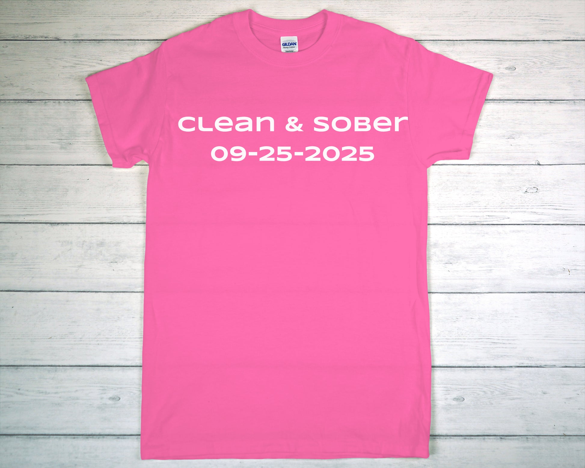 Pink t-shirt with 'clean & sober 09-25-2025' text on a wooden background