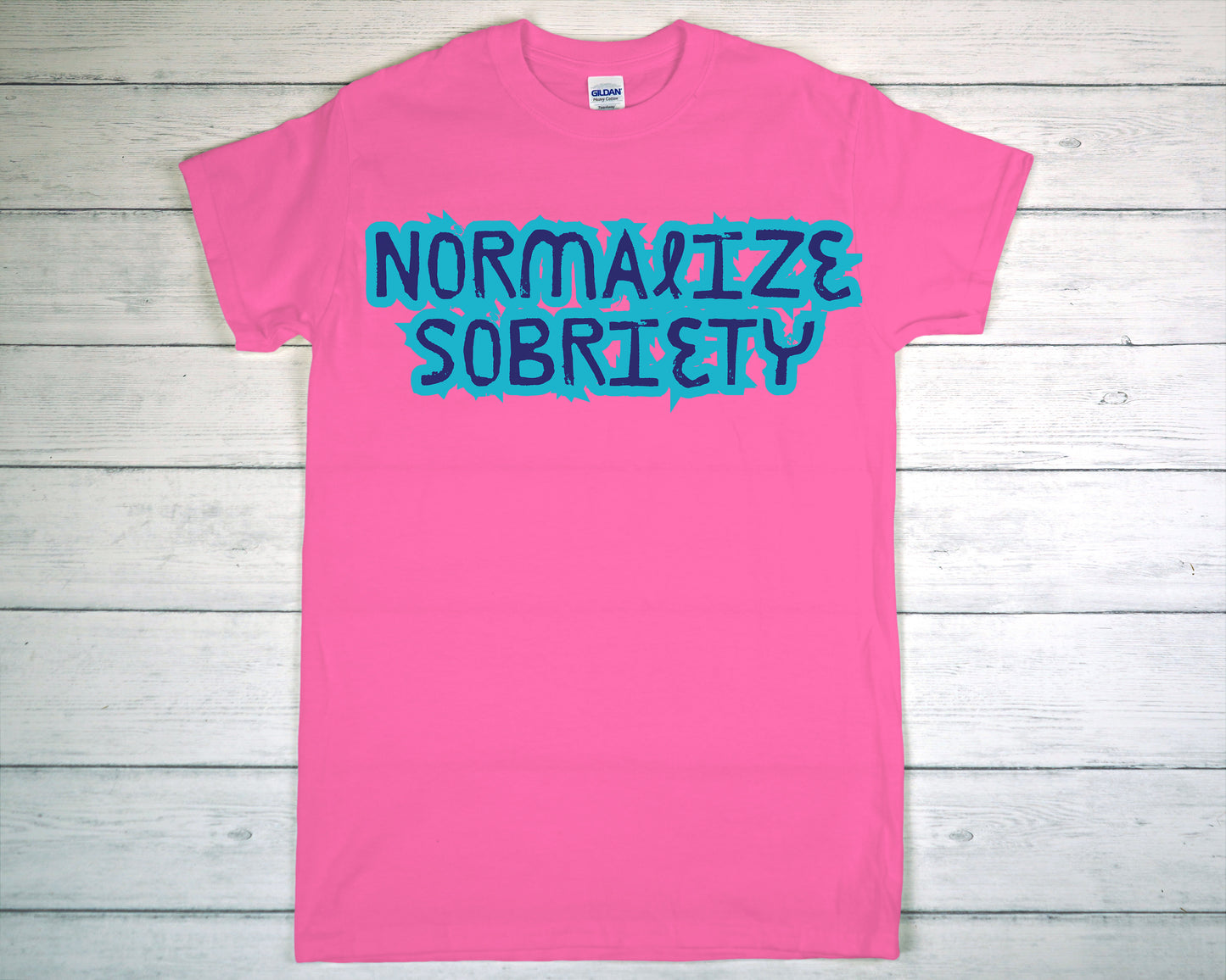 Pink t-shirt with 'Normalize Sobriety' text on a wooden surface