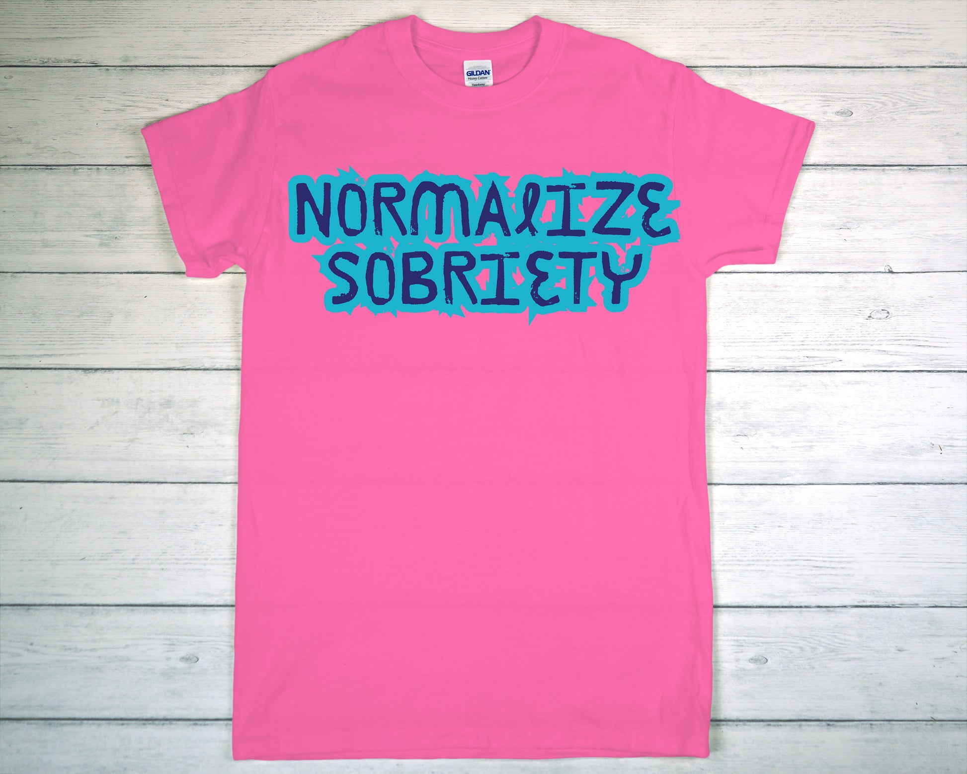 Pink t-shirt with 'Normalize Sobriety' text on a wooden surface