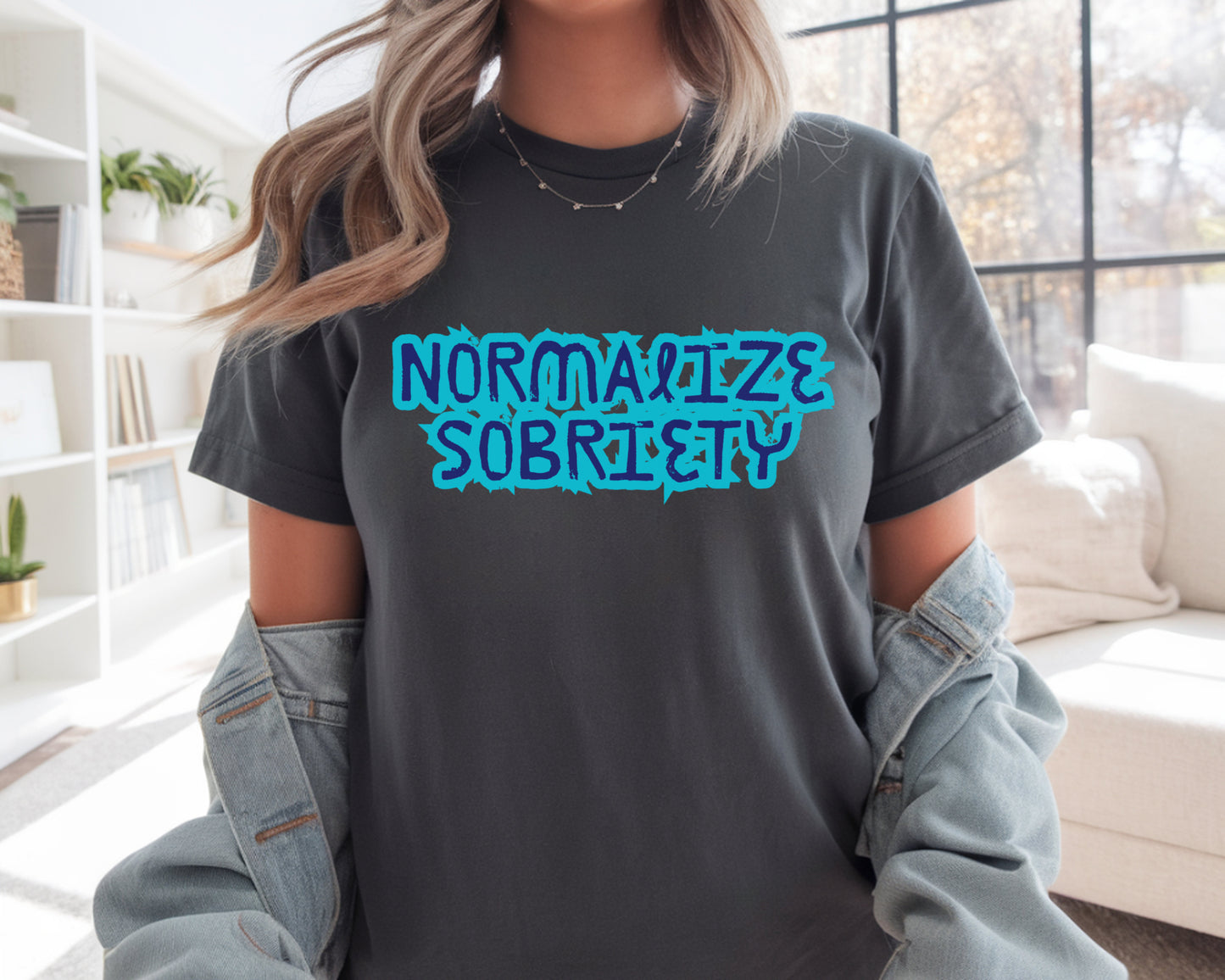 Person wearing a gray t-shirt with 'NORMALIZE SOBRIETY' in blue text, standing in a room with a couch and shelves.