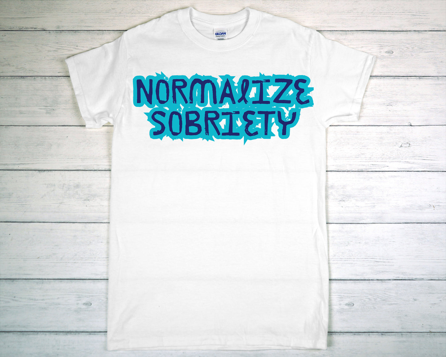 White t-shirt with 'NORMALIZE SOBRIETY' text in blue on a wooden background