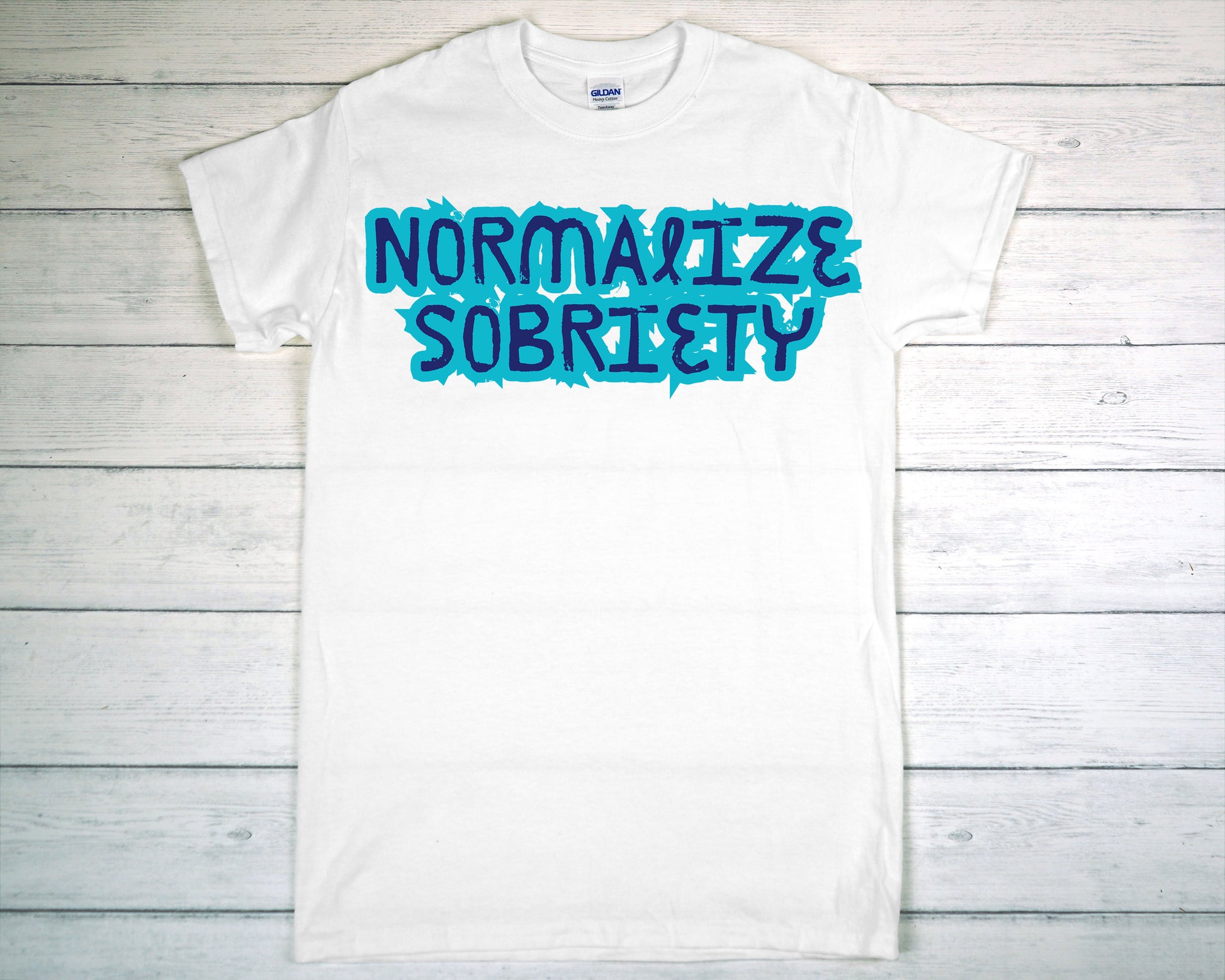 White t-shirt with 'NORMALIZE SOBRIETY' text in blue on a wooden background