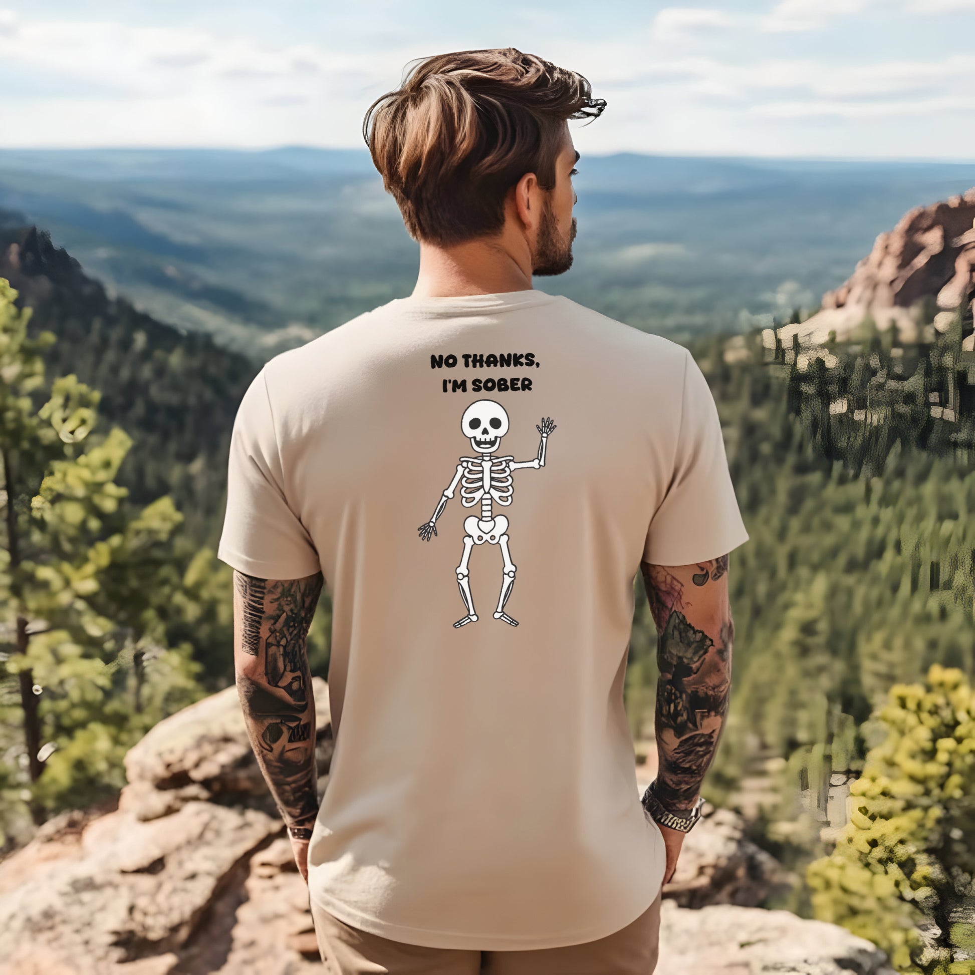 Man wearing a t-shirt with a skeleton design and "No thanks, I'm sober" on the back, standing on a mountain with a scenic view.