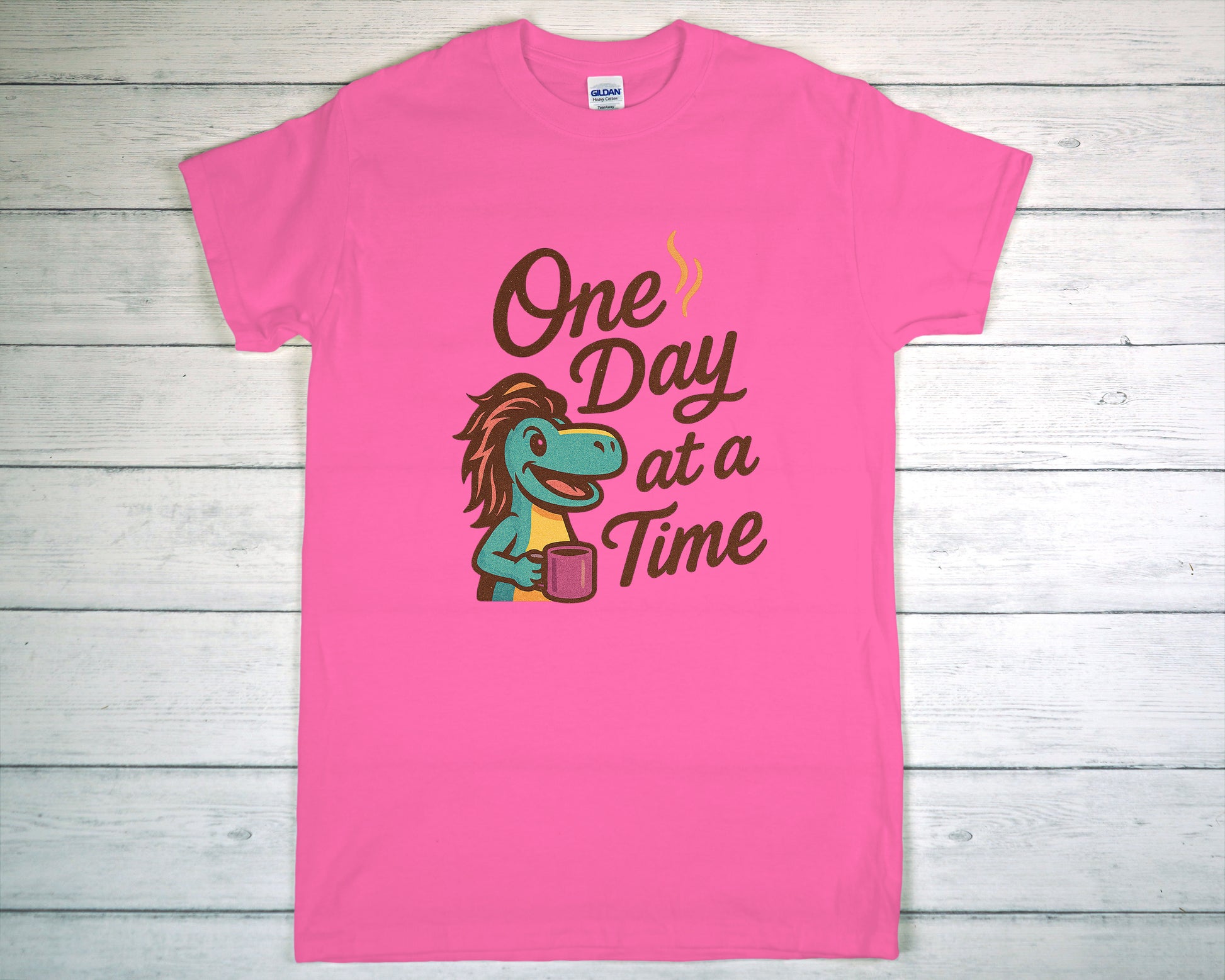 Pink t-shirt with a graphic of a lizard holding a glass and text 'One Day at a Time' on a wooden background