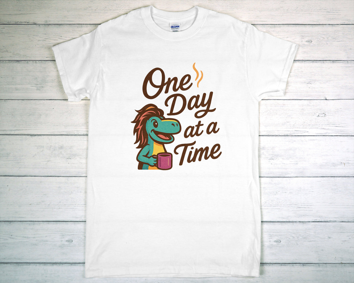 White t-shirt with a cartoon character and 'One Day at a Time' text on a wooden surface