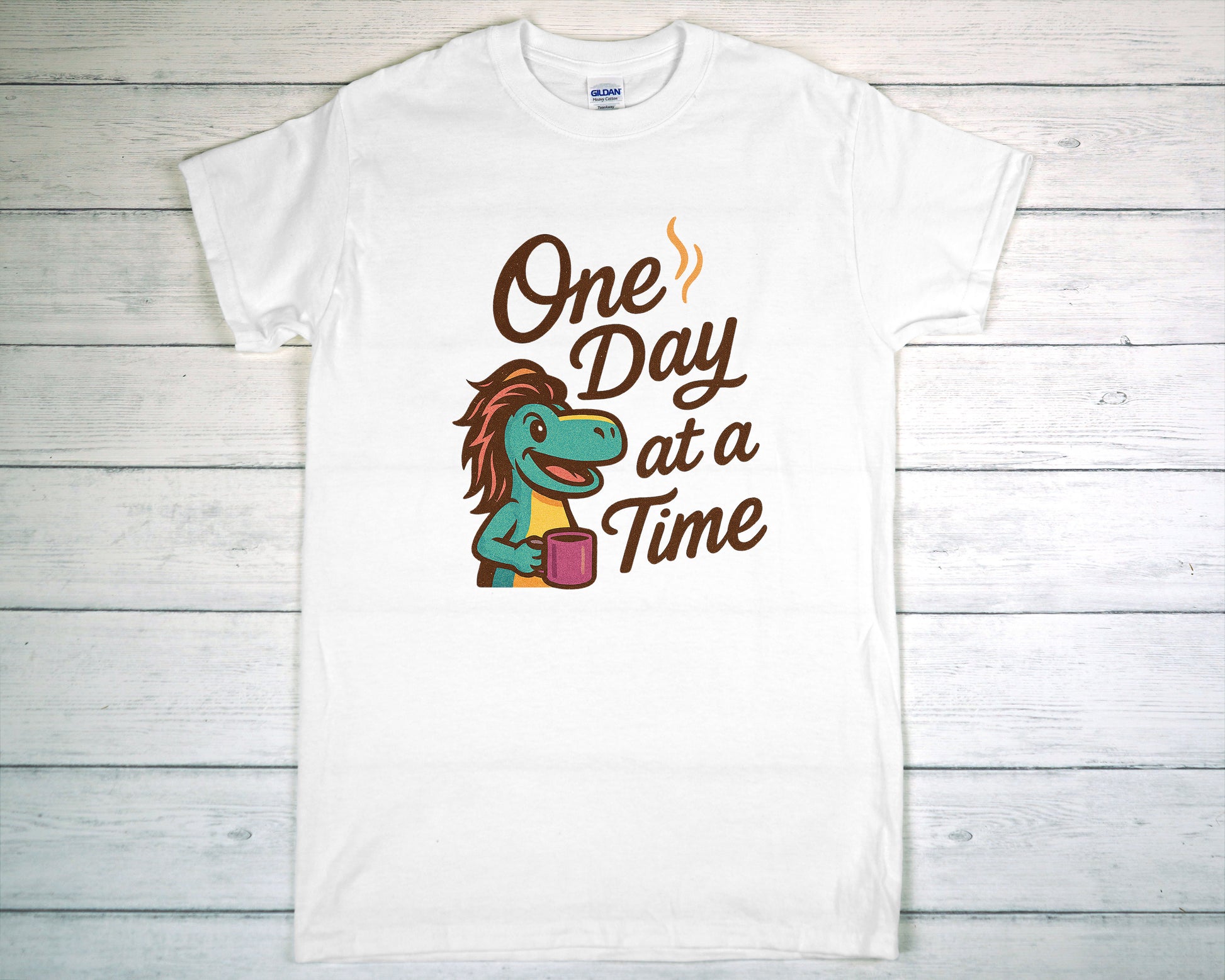 White t-shirt with a cartoon character and 'One Day at a Time' text on a wooden surface