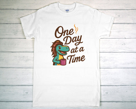 White t-shirt with a cartoon character and 'One Day at a Time' text on a wooden surface