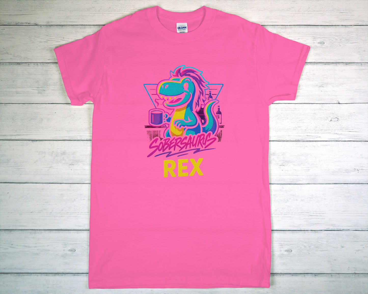 Pink t-shirt with colorful dinosaur graphic and "Sobersaurus Rex" on a wooden surface