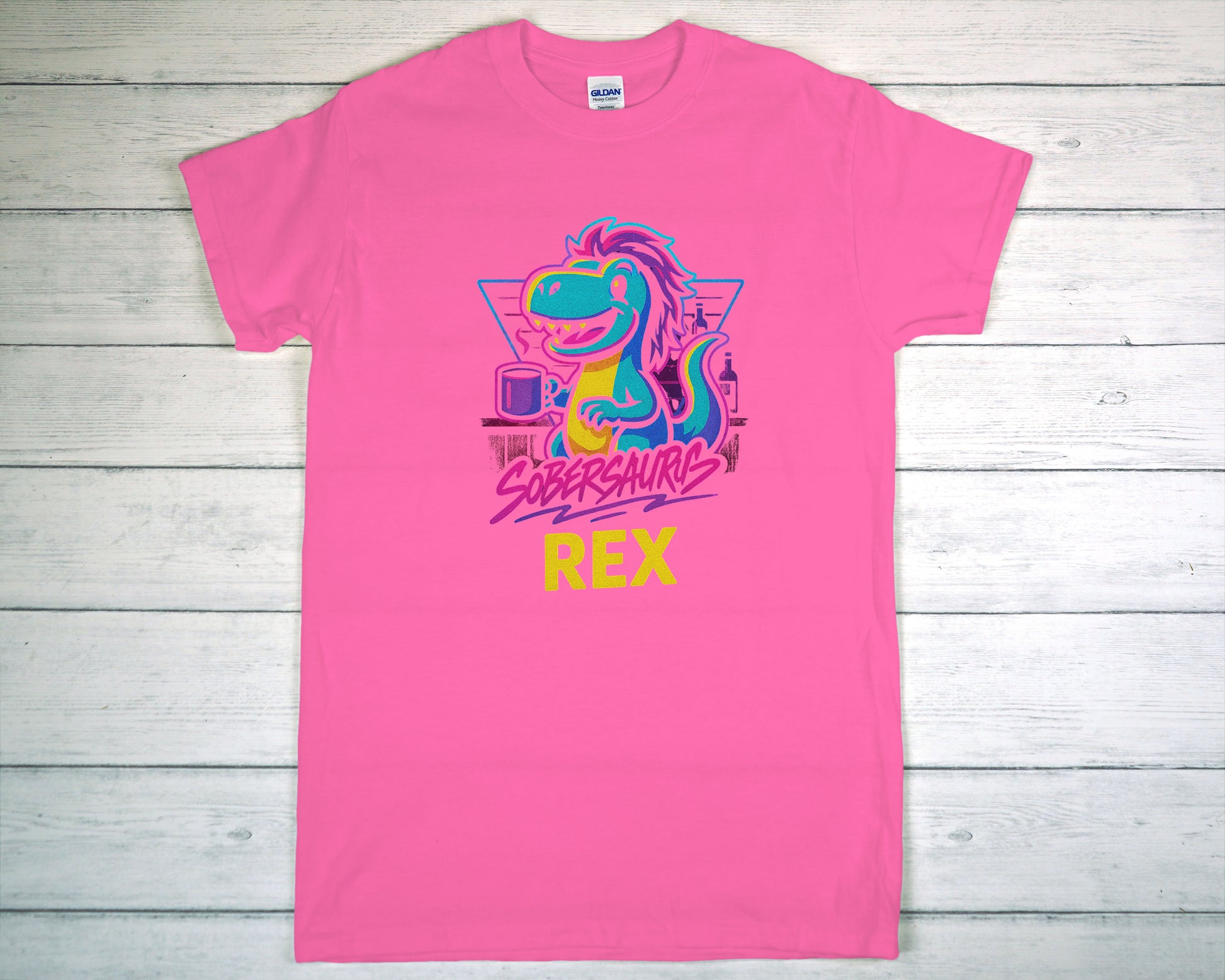 Pink t-shirt with colorful dinosaur graphic and "Sobersaurus Rex" on a wooden surface