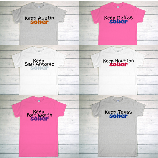 Collection of t-shirts with 'Keep [City] Sober' text on a wooden surface